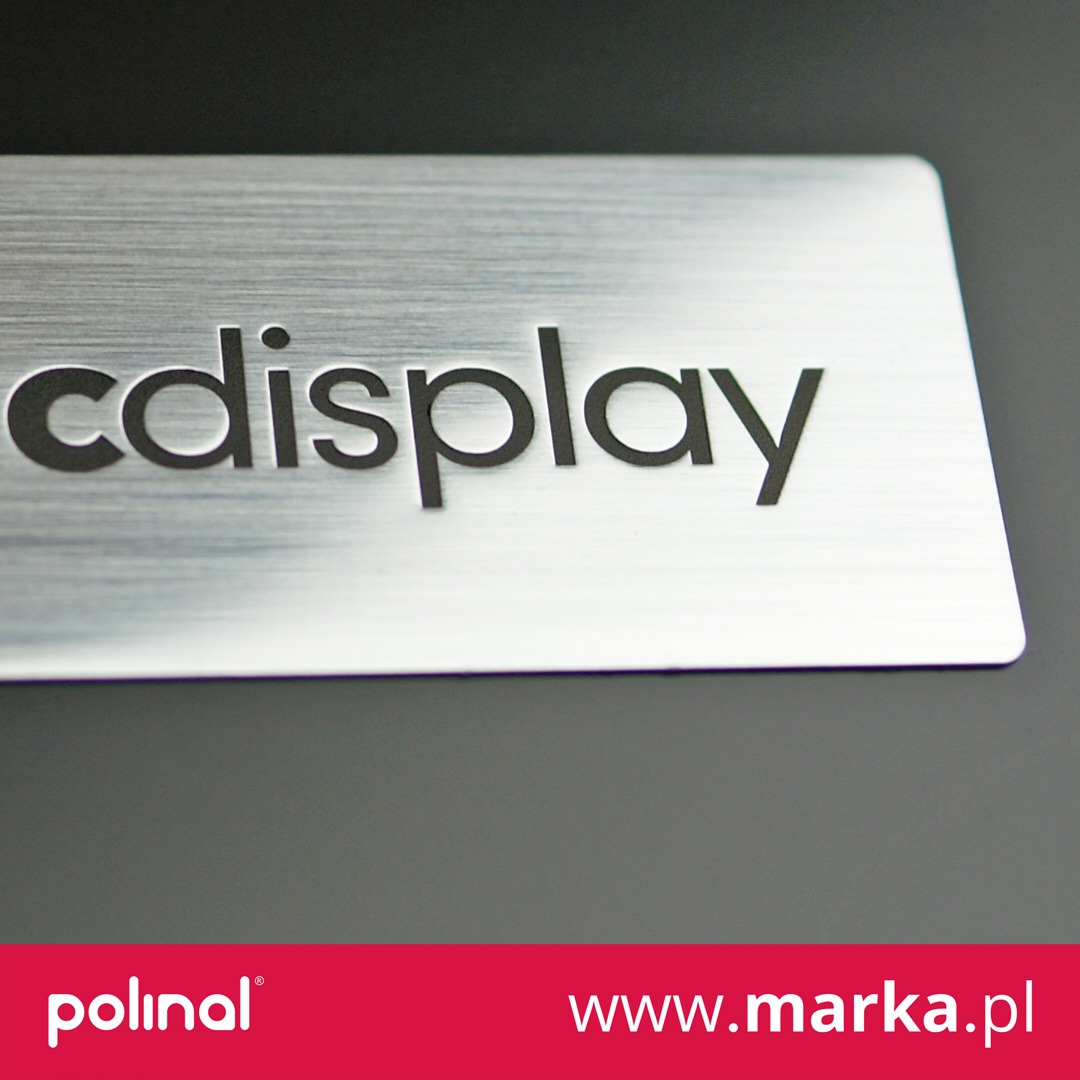 Metalowa naklejka z wygrawerowanym napisem 'cdisplay', wykończenie szczotkowane, umieszczona na ciemnym tle.