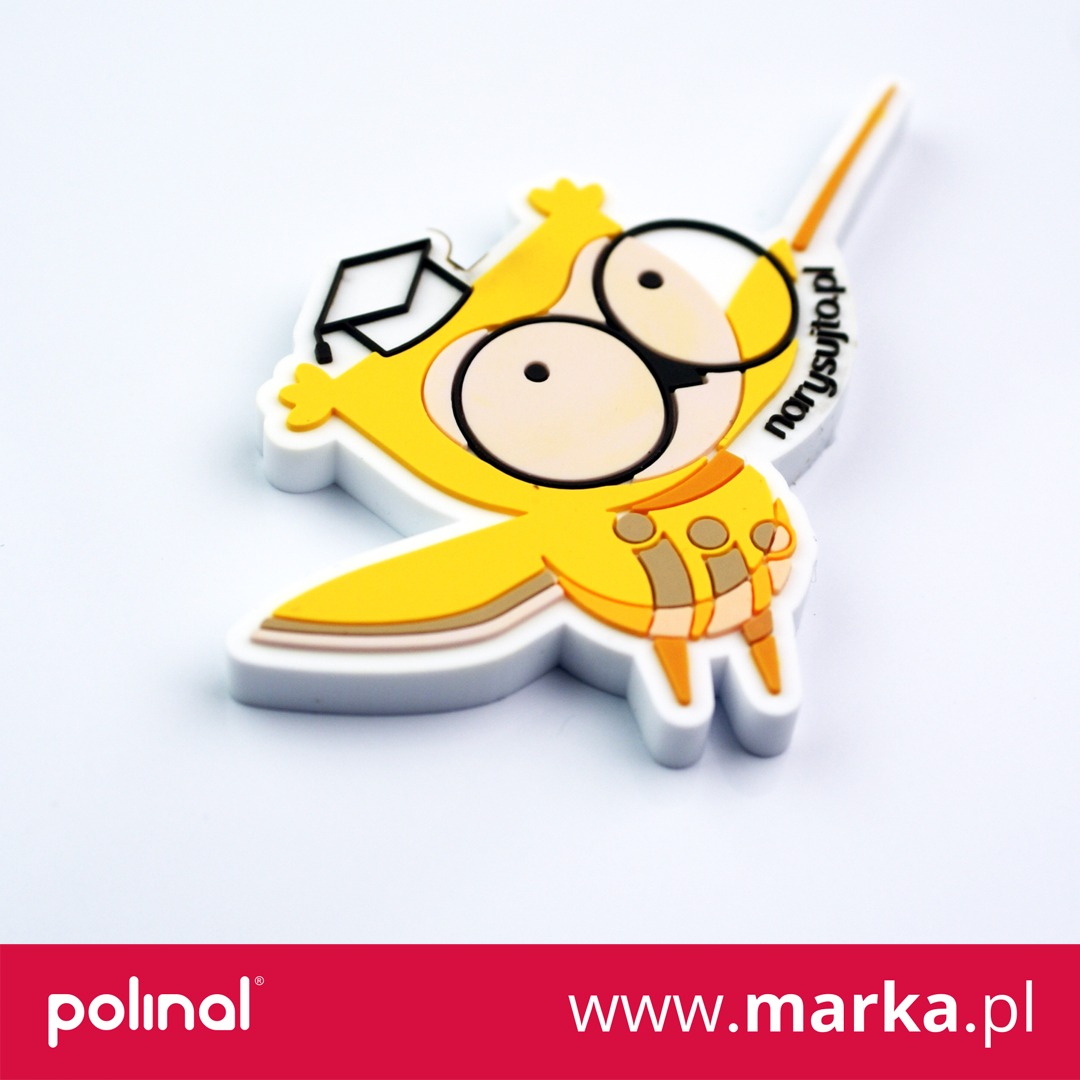 Żółta, gumowa figurka w kształcie sowy z czapką absolwenta, leżąca na białym tle, z nadrukowanym adresem strony internetowej.