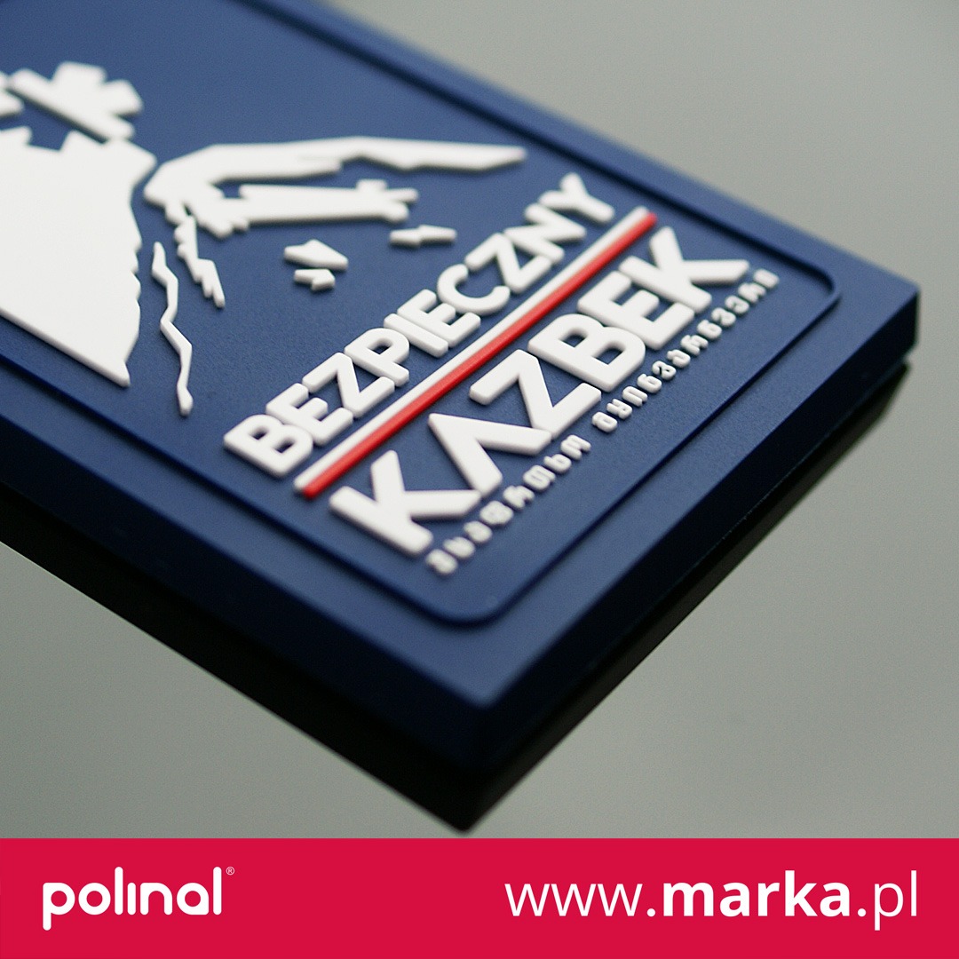 Niebieska, gumowa naszywka z wypukłym białym napisem 'BEZPIECZNY KAZBEK', czerwonym paskiem i białym symbolem graficznym góry.