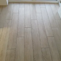 Ułożona podłoga z płytek imitujących drewno w jasnym odcieniu, widok z góry, fuga w kolorze zbliżonym do płytek, przy oknie widoczne gruz i ziemia.