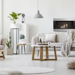 Nowoczesny salon z minimalistycznym kominkiem w zabudowie, szarym dywanem i jasnymi meblami, podkreślony przez duże rośliny doniczkowe.