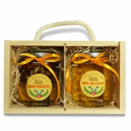 Dwa słoiki miodu (akacjowy i wielokwiatowy z orzechami) w drewnianej skrzynce z jutowym uchwytem, przewiązane pomarańczowymi wstążkami, wypełnione siankiem dekoracyjnym.