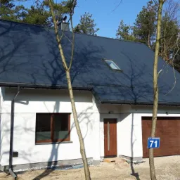 Nowoczesny dom jednorodzinny z ciemnoszarą blachodachówką, białymi ścianami i drewnianą stolarką okienną, otoczony lasem iglastym.