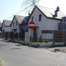 Porin - Szereg identycznych domów jednorodzinnych z szarymi dachami blachodachówkami i białymi elewacjami, oddzielonych drewniano-metalowymi ogrodzeniami, widok z chodnika.