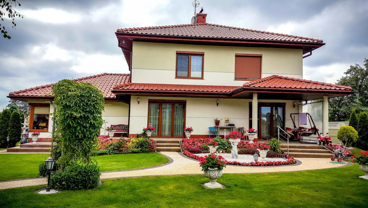 Dwukondygnacyjny dom z czerwonym dachem, zadbanym ogrodem, ścieżką z kostki i dekoracyjnymi donicami z kwiatami.