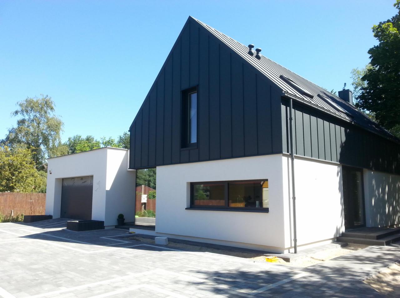 Nowoczesny dom z ciemną, pionowo ułożoną blachą na dachu i górnej części elewacji, białe ściany, szara kostka brukowa na podjeździe, błękitne niebo.