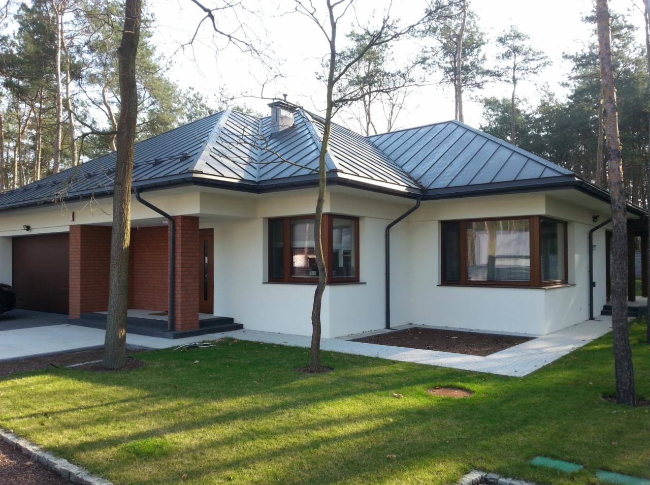 Nowoczesny parterowy dom z ciemnoszarą blachą na dachu, białymi ścianami i brązowymi ramami okiennymi, otoczony zielonym trawnikiem i drzewami.