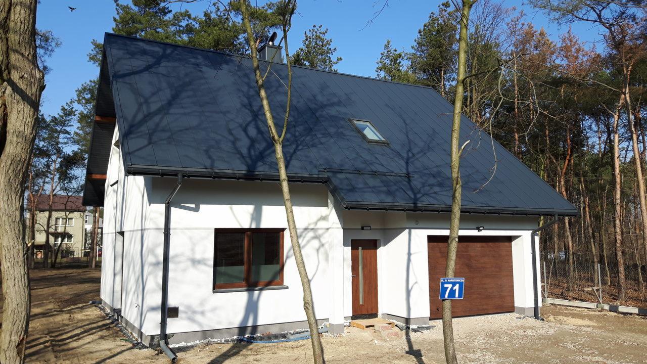 Nowoczesny dom jednorodzinny z ciemnoszarą blachodachówką, białymi ścianami i drewnianą stolarką okienną, otoczony lasem iglastym.