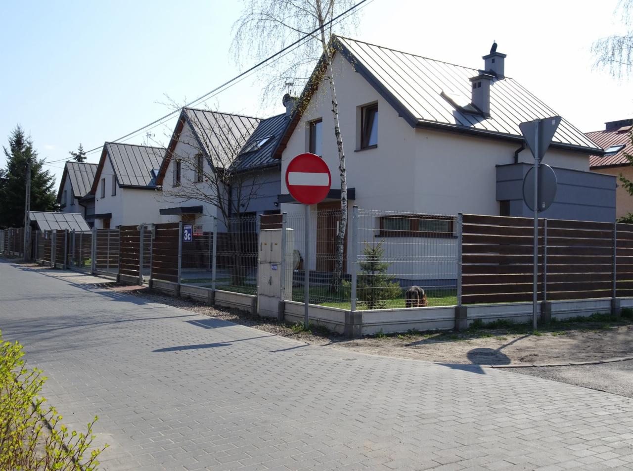 Szereg identycznych domów jednorodzinnych z szarymi dachami blachodachówkami i białymi elewacjami, oddzielonych drewniano-metalowymi ogrodzeniami, widok z chodnika.