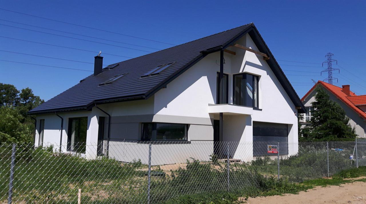 Nowoczesny dom jednorodzinny z ciemną dachówką, białymi ścianami i szarym pasem na elewacji, widoczny przez siatkę ogrodzeniową, na tle błękitnego nieba i linii wysokiego napięcia.