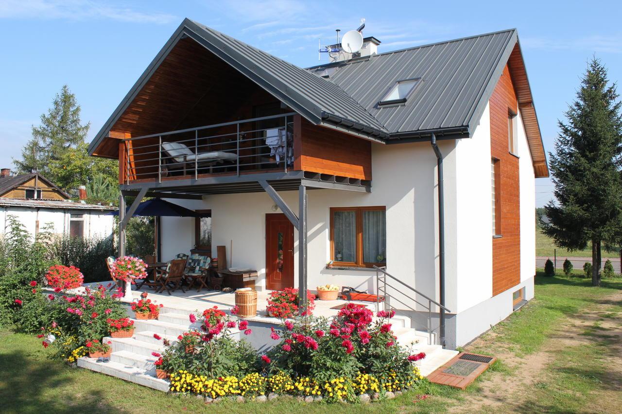 Nowoczesny dom z szarą blachodachówką, drewnianą elewacją i balkonem z metalową balustradą, otoczony ogrodem z kwiatami.