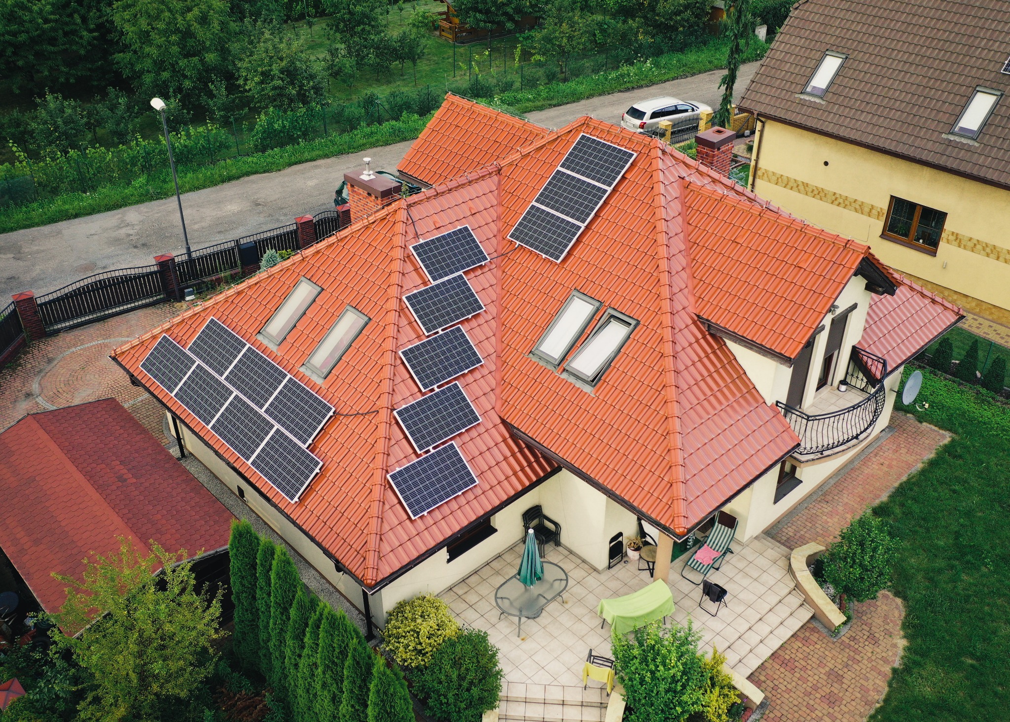 Widok z góry na dom z czerwoną dachówką, na której zamontowane są panele słoneczne, a obok znajduje się taras z meblami ogrodowymi.