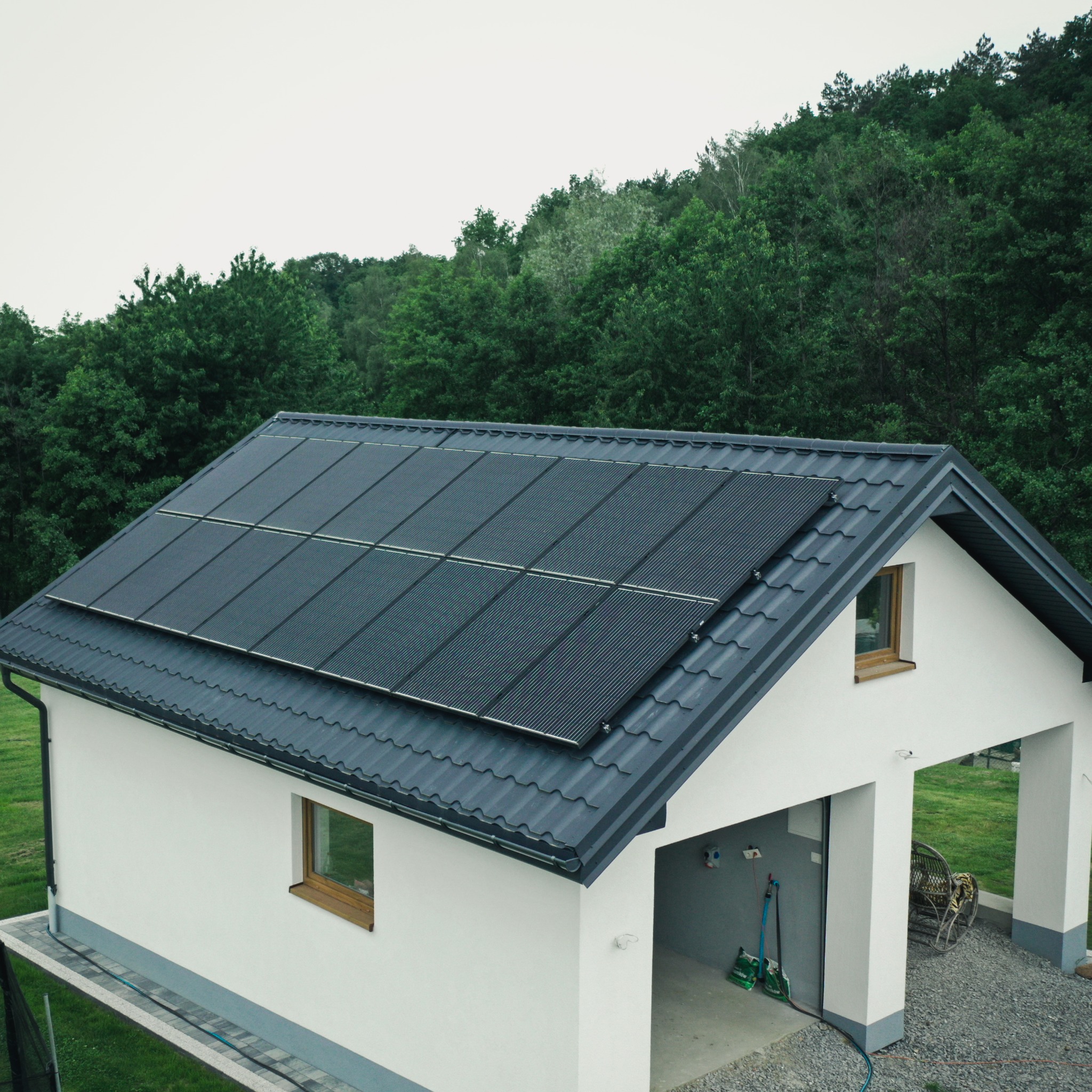 Dom jednorodzinny z panelami słonecznymi na grafitowym dachu, widok z góry, tło stanowią zielone drzewa.