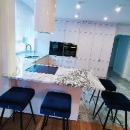 Nowoczesna kuchnia z białymi szafkami, wyspą z blatem o wzorze marmuru i czterema granatowymi hokerami z czarnymi nogami.