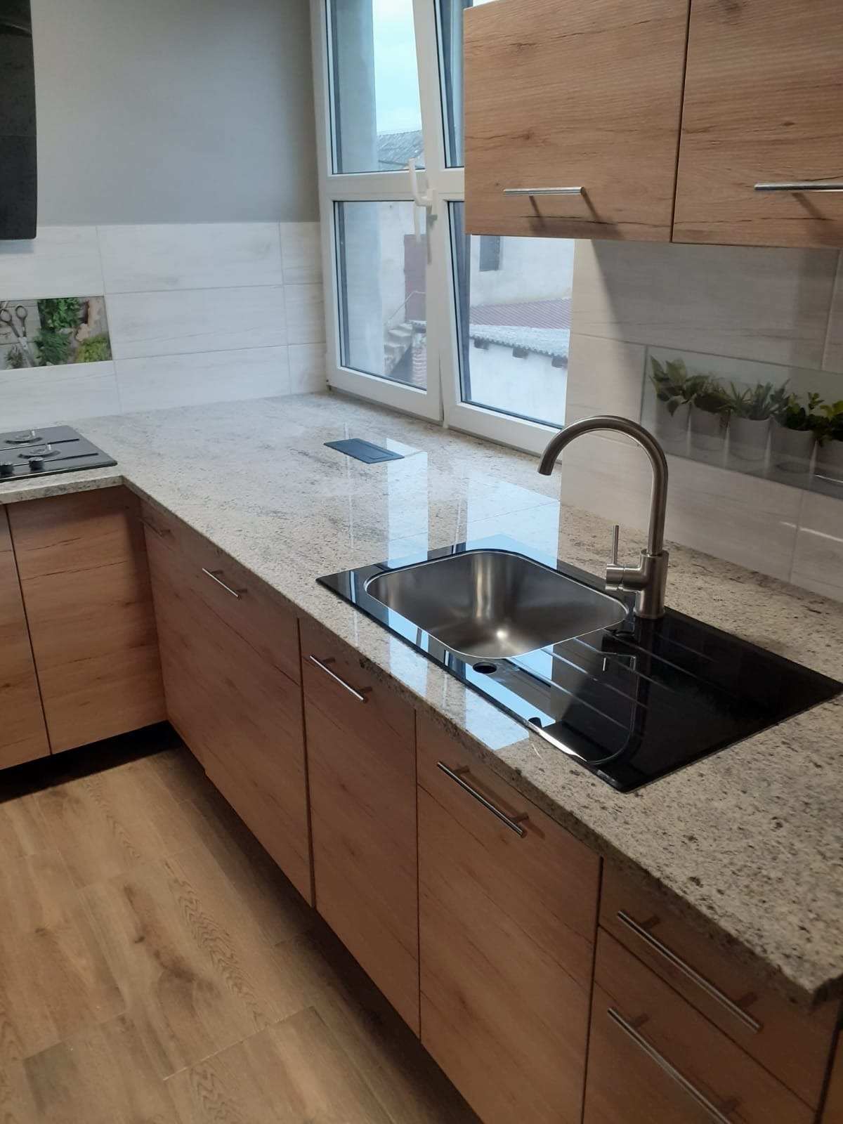 Jasny blat z drobnym wzorem kamienia, zintegrowany z czarnym zlewem i płytą grzewczą, drewniane szafki kuchenne, okno w tle.