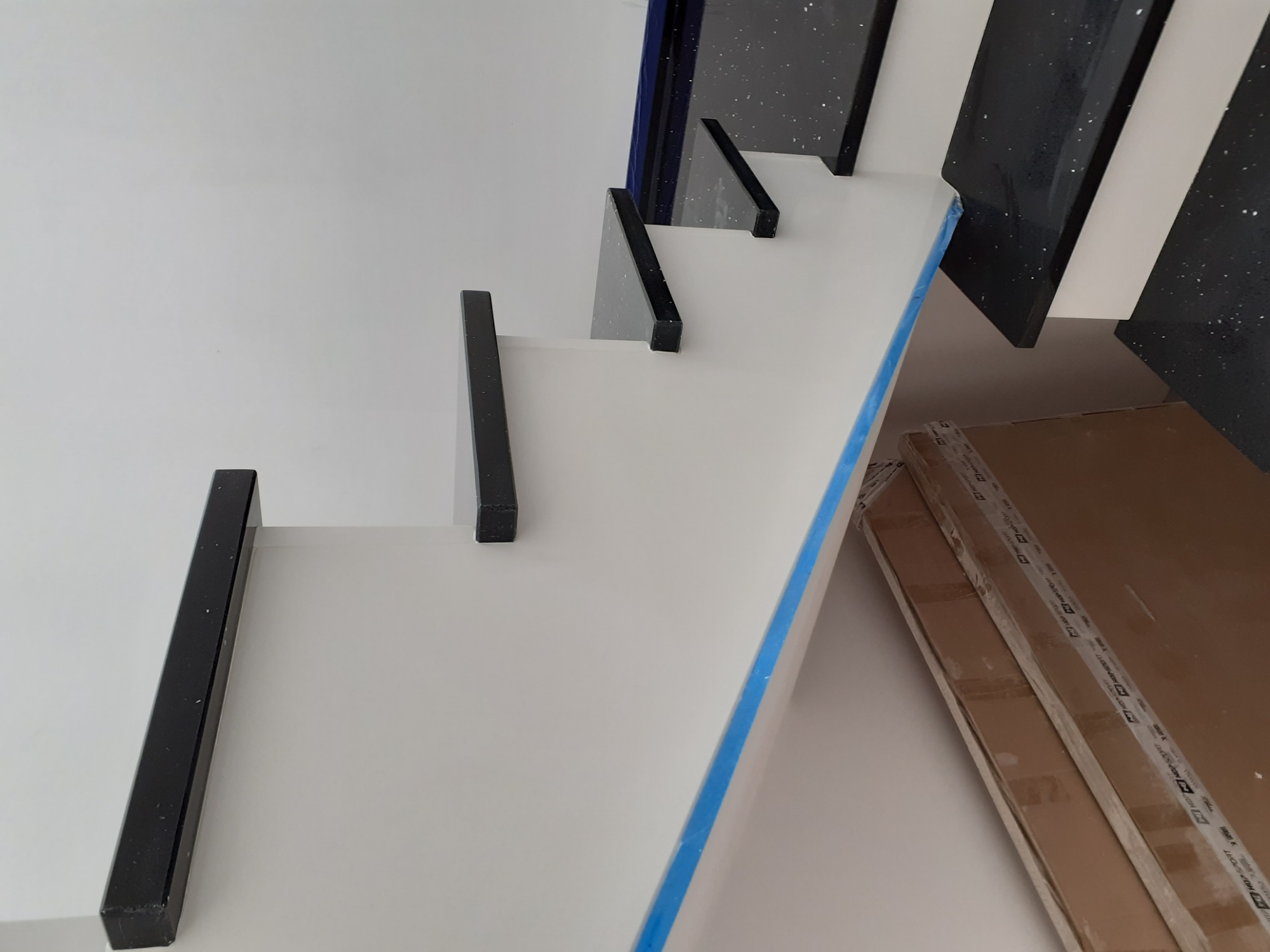 Białe schody z czarnymi elementami, zabezpieczone niebieską folią, widoczne kartony w tle.