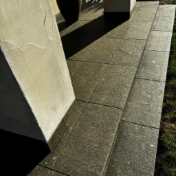 Ułożona nawierzchnia z ciemnej, ziarnistej kostki na schodach wejściowych, z widocznym fragmentem kolumny i donicy z rośliną w tle.