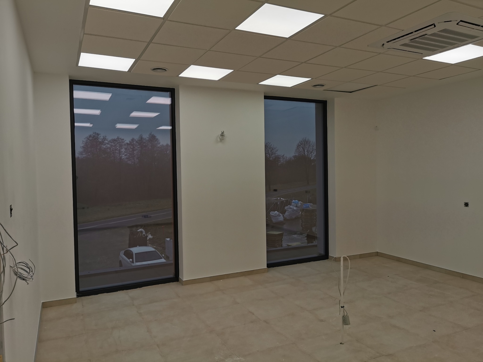 Wnętrze pustego pomieszczenia z dużymi oknami, widoczne kable wystające ze ściany, przygotowane do wykończenia.