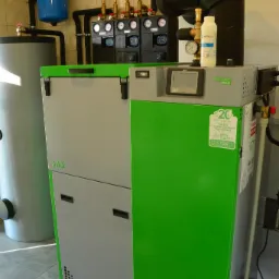 Kocioł SAS Bio EFEKT Plus w kotłowni z szarymi płytkami podłogowymi. Widoczne elementy instalacji i zbiorniki. Urządzenie z zielonym panelem i czarnymi detalami.