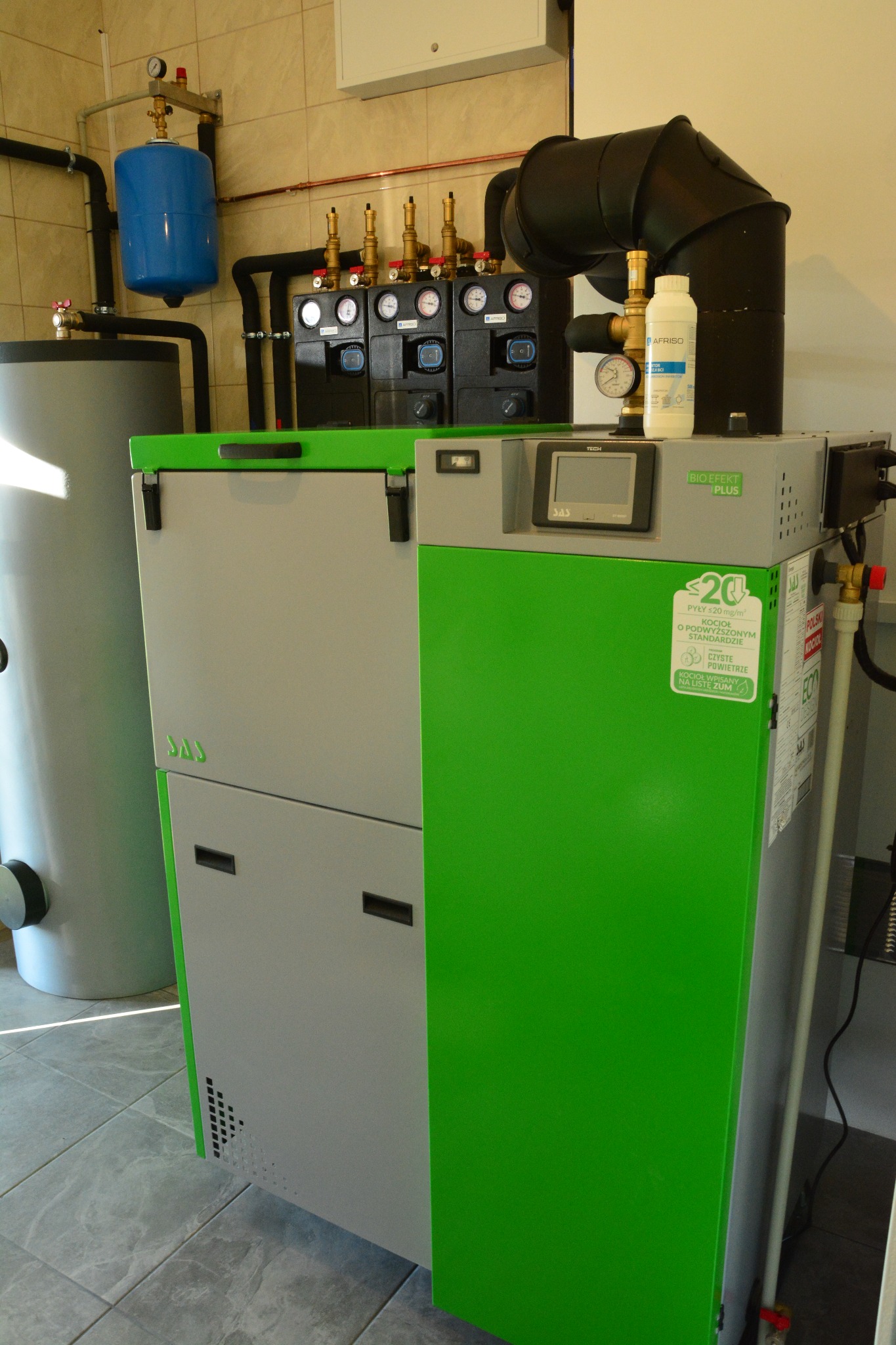 Kocioł SAS Bio EFEKT Plus w kotłowni z szarymi płytkami podłogowymi. Widoczne elementy instalacji i zbiorniki. Urządzenie z zielonym panelem i czarnymi detalami.