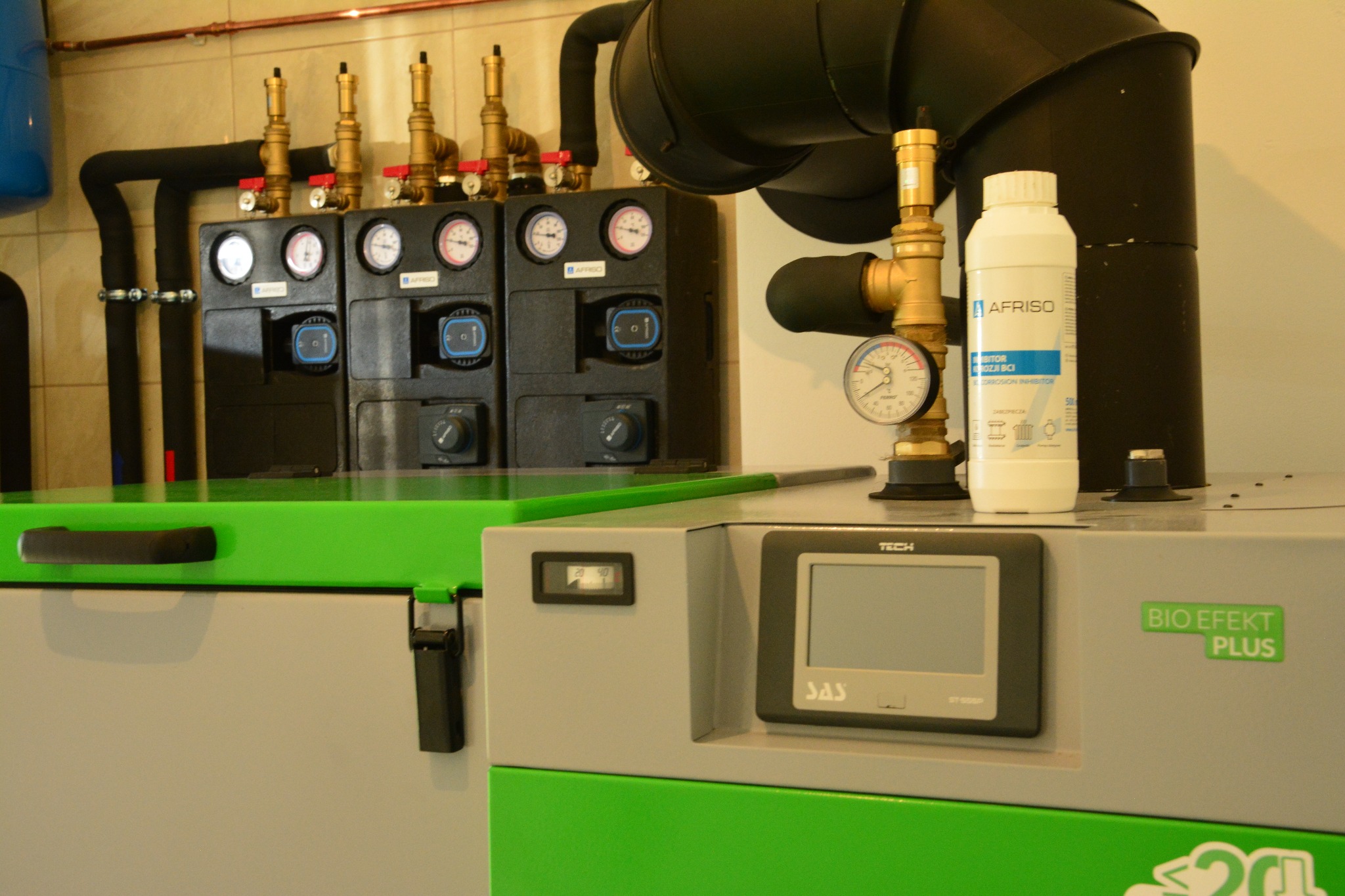 Instalacja kotła Bio Efekt Plus z czarnymi rurami, manometrami Afriso i białym pojemnikiem z napisem Afriso, widoczne elementy sterujące Tech SAS.