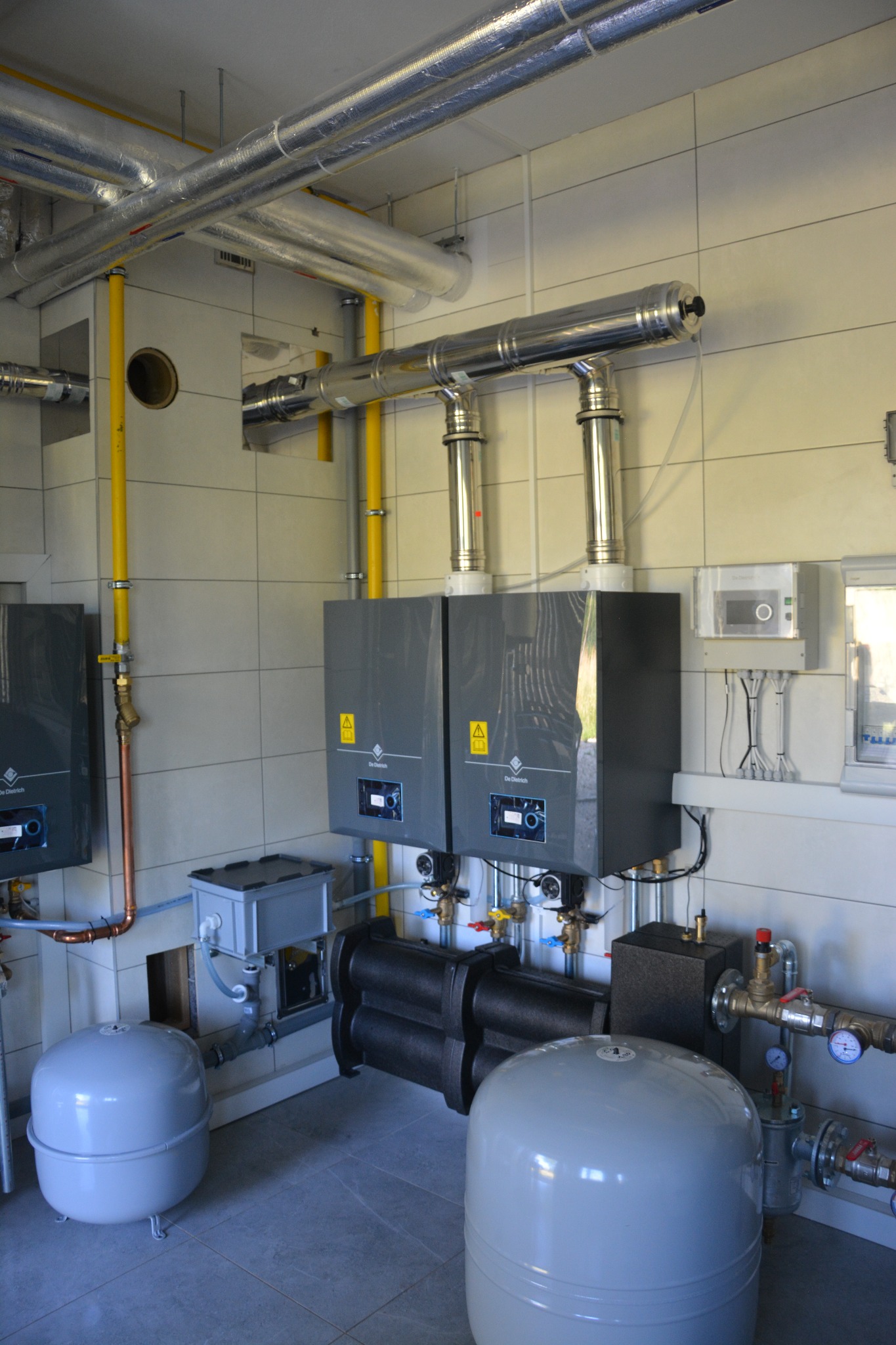 Zakończona instalacja grzewcza w kotłowni: dwa kotły De Dietrich, orurowanie, zbiorniki wyrównawcze, manometry, zawory i  żółte rury gazowe przy ścianie z płytek.