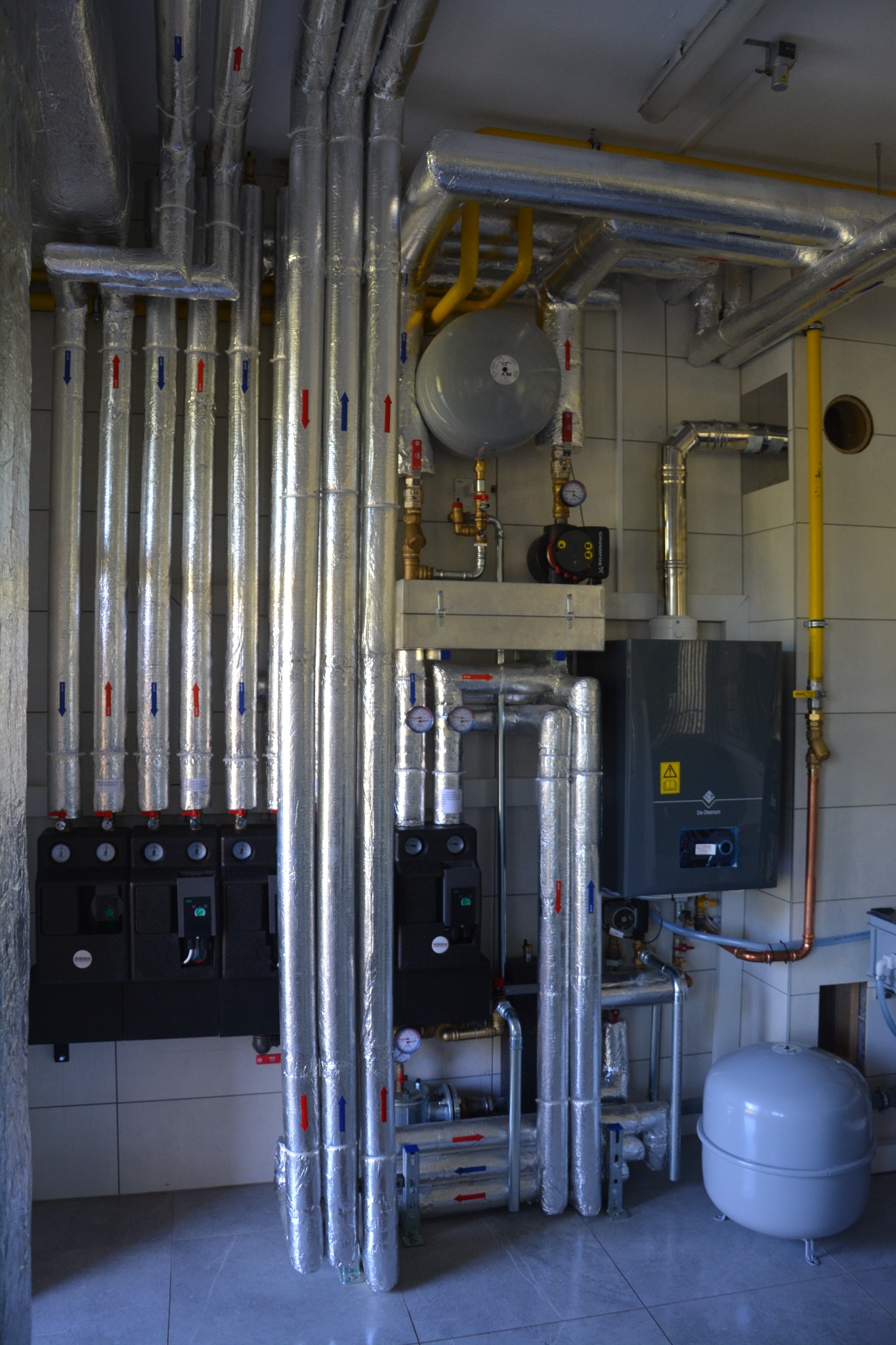 Kompleksowa instalacja grzewcza: rury z izolacją, czarne skrzynki rozdzielcze, szary piec gazowy De Dietrich, zbiornik wyrównawczy i żółta rura gazowa na tle białych płytek.