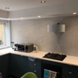 Nowoczesna kuchnia z szarymi szafkami, białym blatem i ścianą wyłożoną mozaiką w drobne biało-czarne kropki. Widoczny sprzęt AGD: mikrofalówka i płyta grzewcza. Nad płytą minimalistyczny okap.