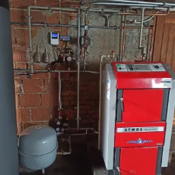 Czerwono-szary piec na paliwo stałe Atmos Generator DC 20 GS z podłączoną instalacją CO, w tym zbiornik wyrównawczy i liczne rury miedziane na tle ściany z czerwonej cegły i białym sterownikiem TECH.