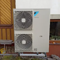 Zewnętrzna jednostka pompy ciepła Daikin Altherma zamontowana na metalowej konstrukcji przy budynku z żółtą drewnianą elewacją i fragmentem okna z roślinami doniczkowymi na parapecie.