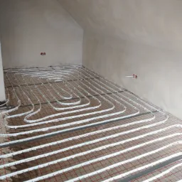 Białe rurki ogrzewania podłogowego ułożone na folii izolacyjnej z czerwoną siatką w surowym wnętrzu poddasza ze ścianami w szarym tynku.