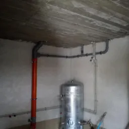 Surowy betonowy strop z instalacją hydrauliczną, w tym pomarańczowa rura pionowa i srebrny zbiornik akumulacyjny w rogu pomieszczenia w stanie deweloperskim.