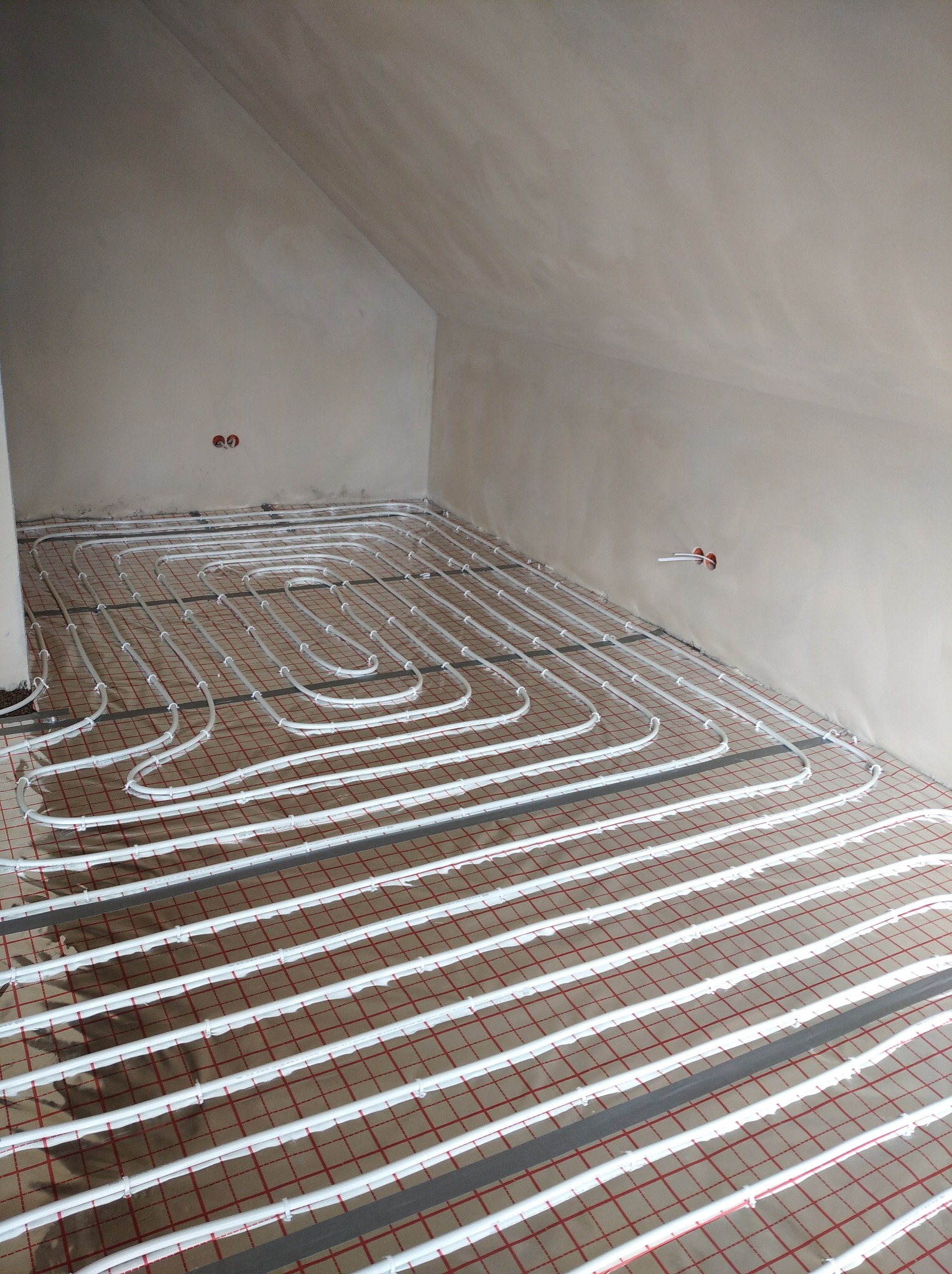 Białe rurki ogrzewania podłogowego ułożone na folii izolacyjnej z czerwoną siatką w surowym wnętrzu poddasza ze ścianami w szarym tynku.