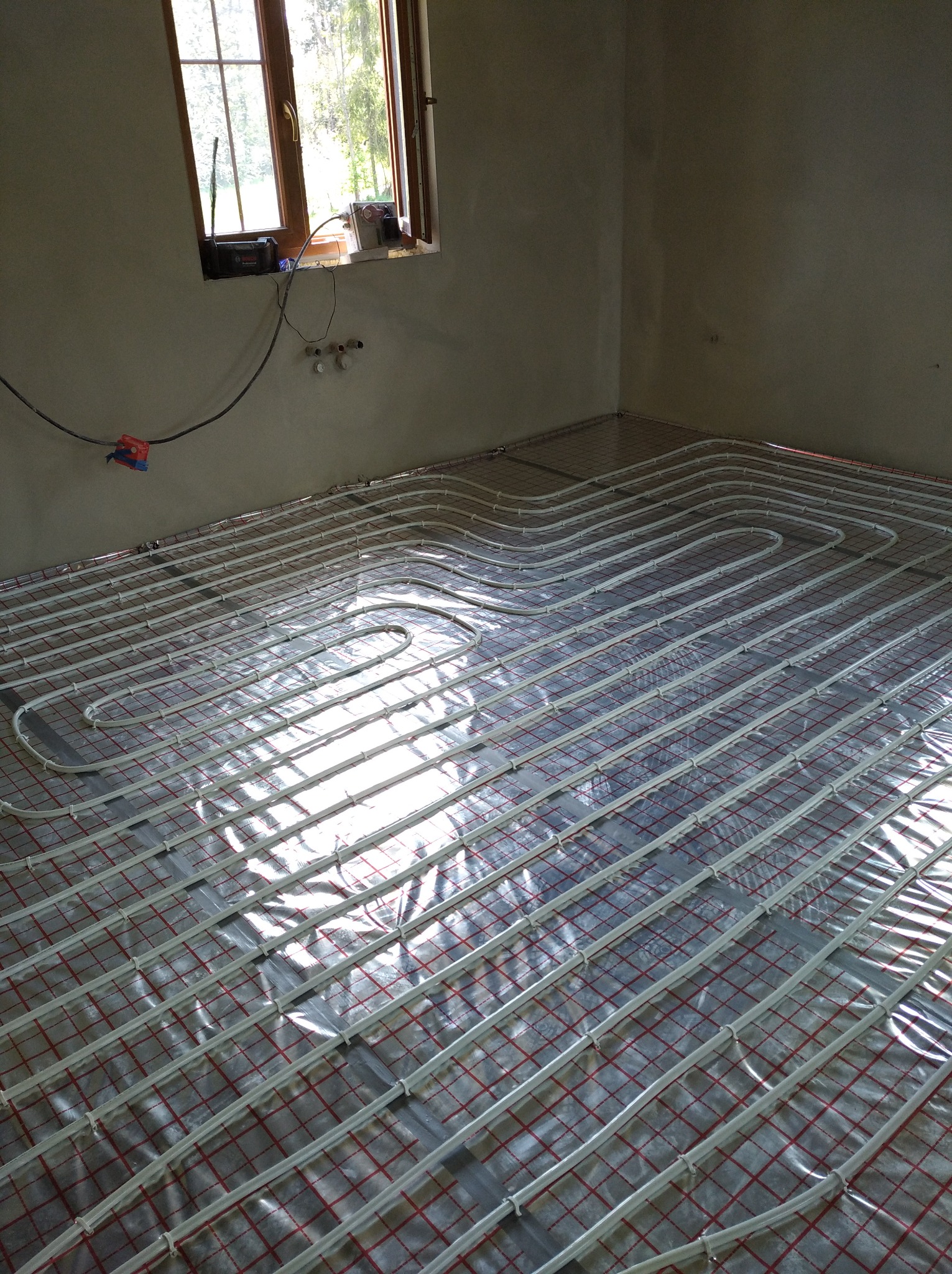 Rury ogrzewania podłogowego ułożone na folii izolacyjnej z czerwoną siatką, widok z góry na surowy stan posadzki w pokoju z oknem.