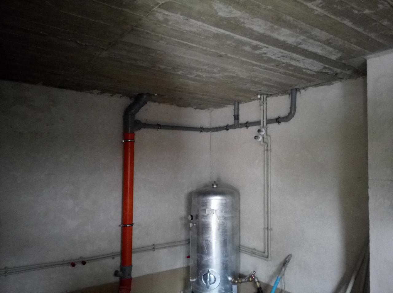 Surowy betonowy strop z instalacją hydrauliczną, w tym pomarańczowa rura pionowa i srebrny zbiornik akumulacyjny w rogu pomieszczenia w stanie deweloperskim.