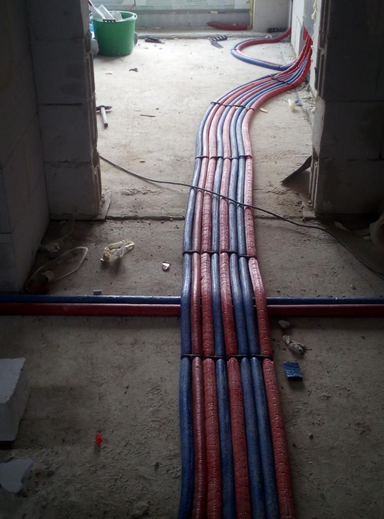 Równolegle ułożone, izolowane rury grzewcze (czerwone i niebieskie) prowadzone przez surowy otwór drzwiowy w trakcie prac instalacyjnych. Widoczne elementy konstrukcyjne z betonu komórkowego...