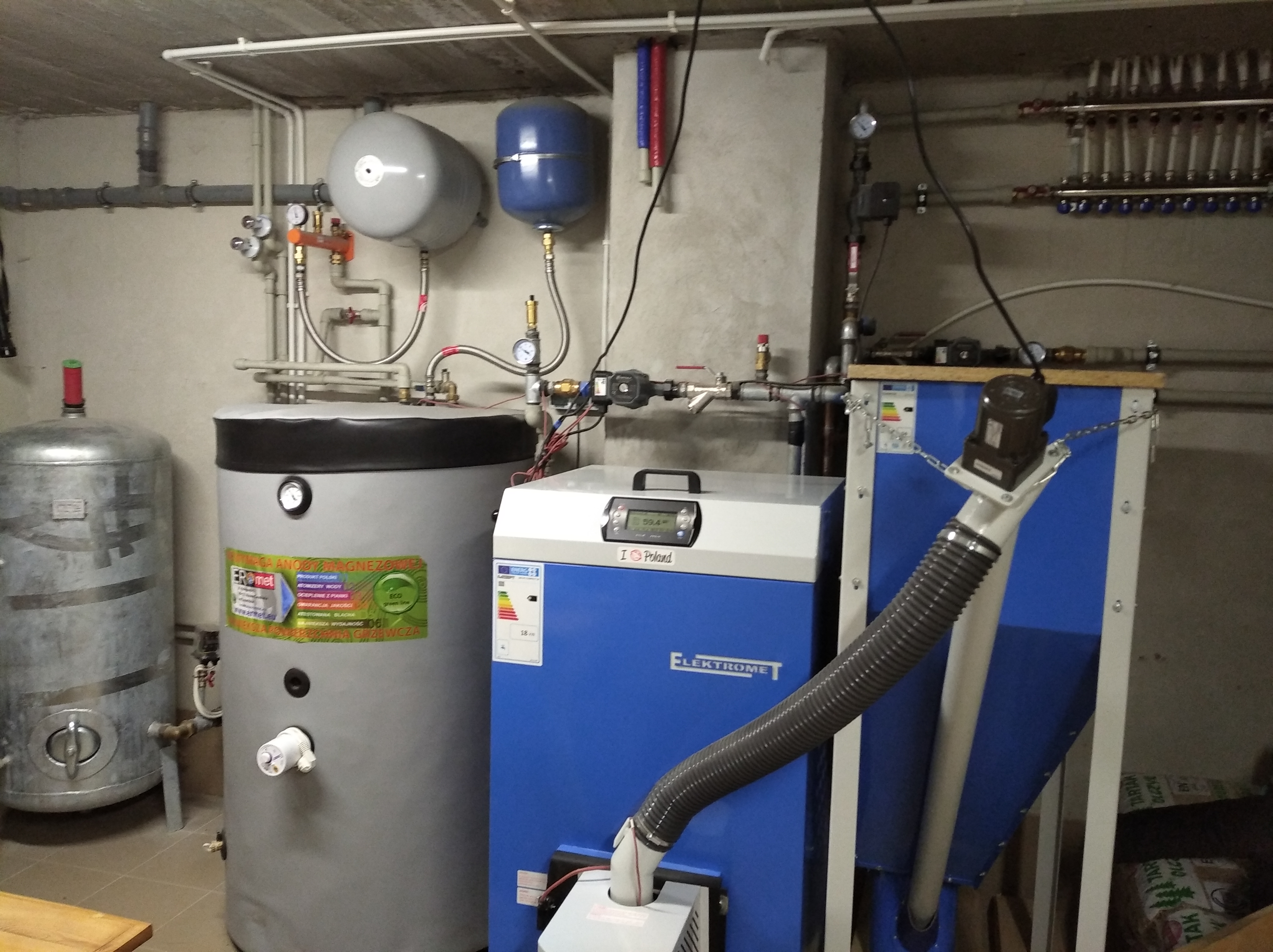 Zestaw urządzeń grzewczych w kotłowni: zbiornik buforowy, zasobnik CWU z naklejką producenta Eromet, niebieski piec na paliwo stałe Elektromet z podajnikiem, manometry i rury instalacji grzewczej.