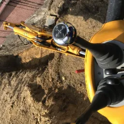 Żółta mini koparka z widocznymi dźwigniami sterującymi i wykopem w ziemi w tle, prawdopodobnie prace ziemne.