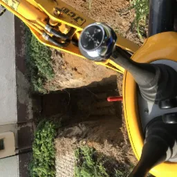 Żółta koparka JCB częściowo widoczna nad wykopem w ziemi, z widocznymi elementami sterującymi i fragmentem budynku w tle.