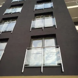 Nowoczesny budynek z ciemną elewacją i balkonami z przezroczystymi szklanymi panelami oraz metalowymi poręczami, uchwycony pod kątem z dołu.