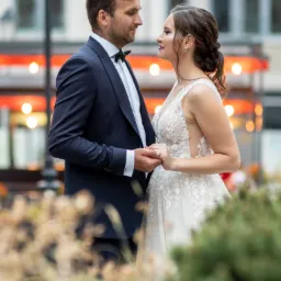 Para młoda trzymająca się za ręce, ona w koronkowej sukni z głębokim dekoltem, on w granatowym smokingu z muchą, zapatrzeni w siebie na tle miejskiej architektury z dekoracyjnym oświetleniem.