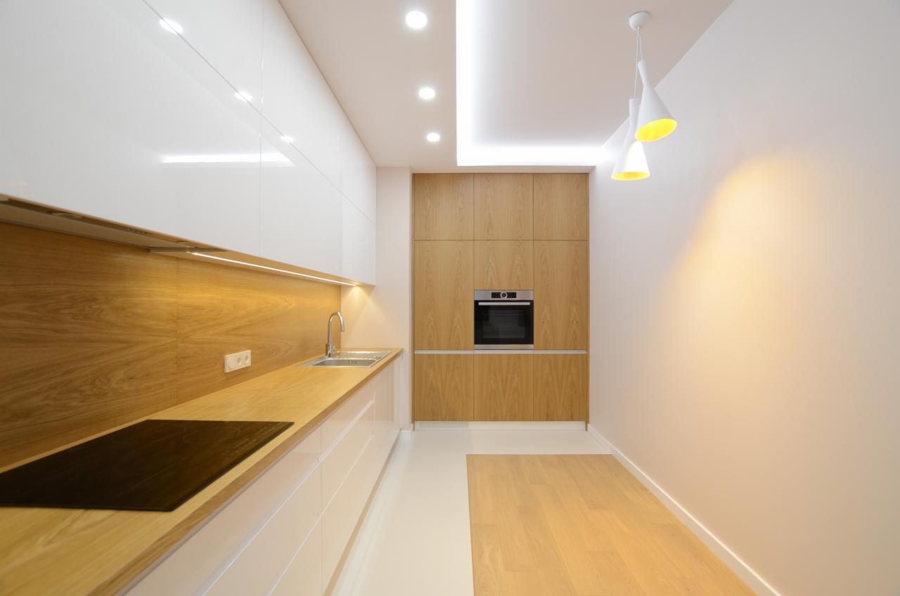 Nowoczesna kuchnia z białymi szafkami dolnymi i górnymi, drewnianym blatem z wbudowaną płytą indukcyjną, oraz wysoką zabudową z forniru dębowego z wbudowanym piekarnikiem, oświetlona trzema...