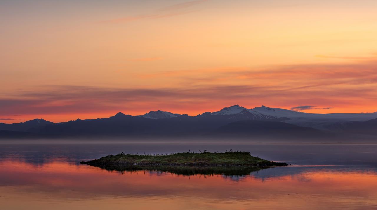 Malownicza wyspa z zielenią na spokojnym jeziorze o zachodzie słońca, z ciemnymi górami w tle i odbiciem w wodzie.