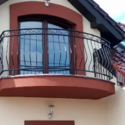 Czarna, metalowa balustrada balkonowa z dekoracyjnymi elementami na tle jasnej elewacji domu z brązowymi akcentami i czerwonym dachem.