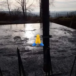 Fundament pod dom w trakcie wylewania betonu, widoczna stalowa siatka zbrojeniowa i rura do pompowania betonu, w tle krajobraz.