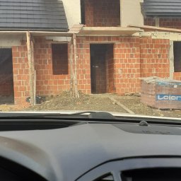 Domel - Surowy stan budowy domu z czerwonej cegły widziany z wnętrza samochodu, widoczne niezamknięte okna i drzwi, na dachu czarna dachówka.