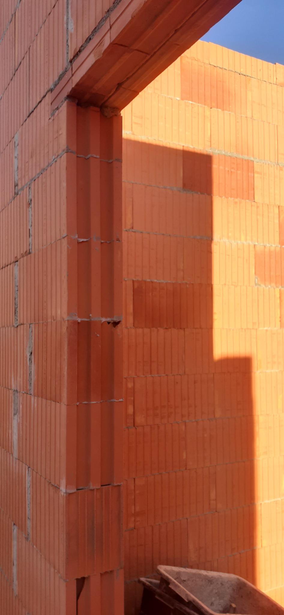 Wznoszenie muru z czerwonych bloczków ceramicznych, widoczny narożnik i nadproże otworu okiennego lub drzwiowego, w tle niebieskie niebo.