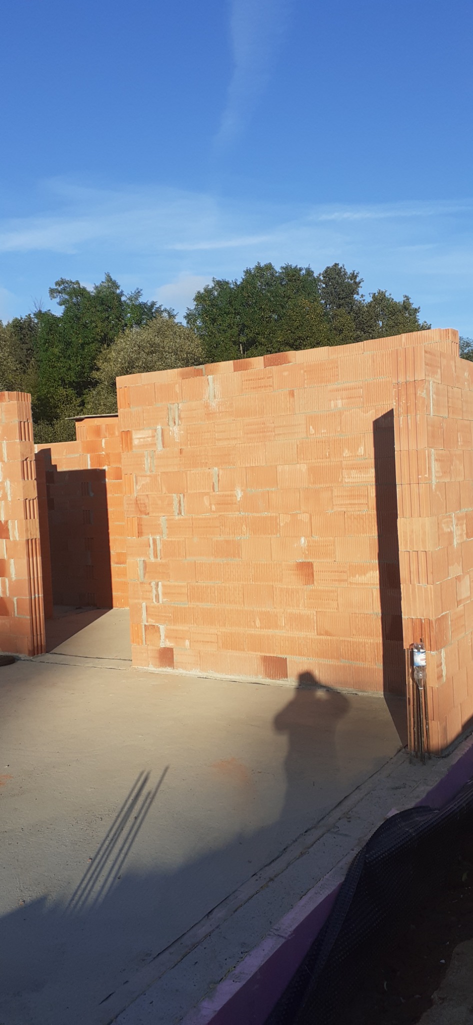 Wznoszone ściany parteru z czerwonej cegły ceramicznej na wylewce fundamentowej, widoczne zarysy przyszłych pomieszczeń, w tle zieleń drzew i błękitne niebo, cień osoby fotografującej na posadzce.