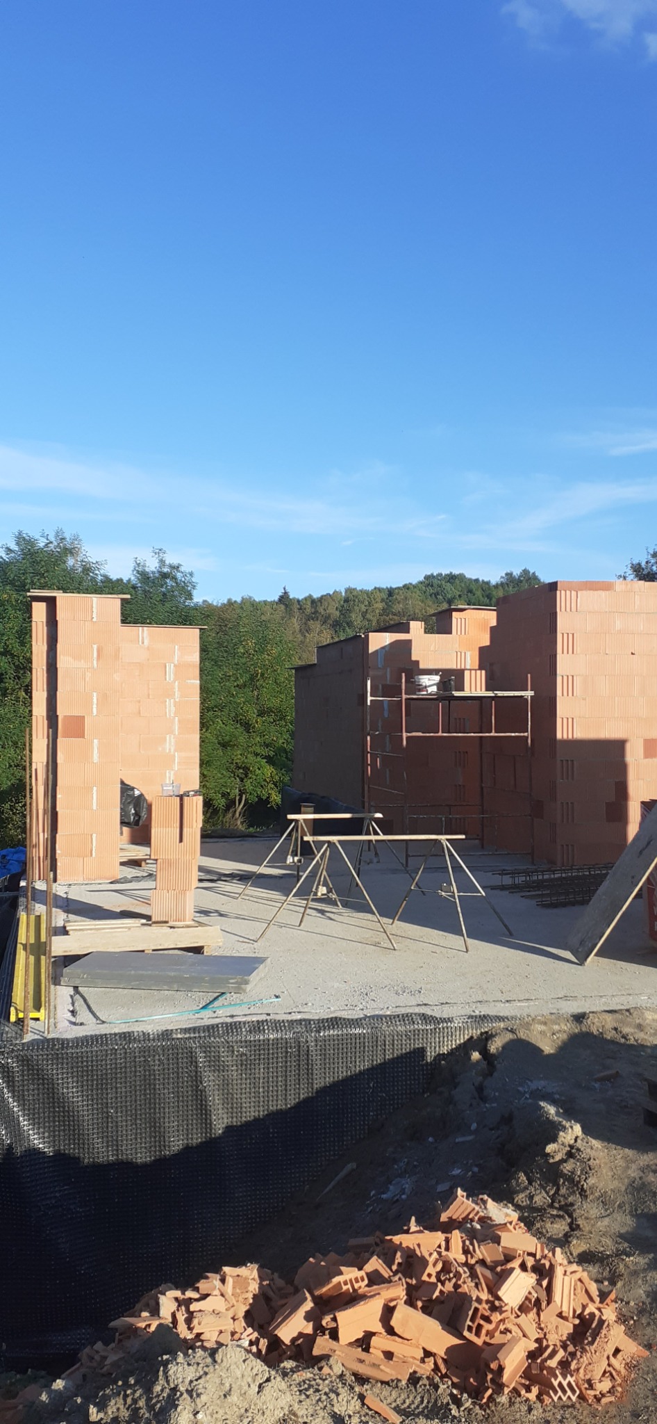 Wznoszone ściany domu z czerwonej cegły ceramicznej, widoczne rusztowania i sterty gruzu ceglanego na placu budowy, tło stanowi zieleń drzew i błękitne niebo.