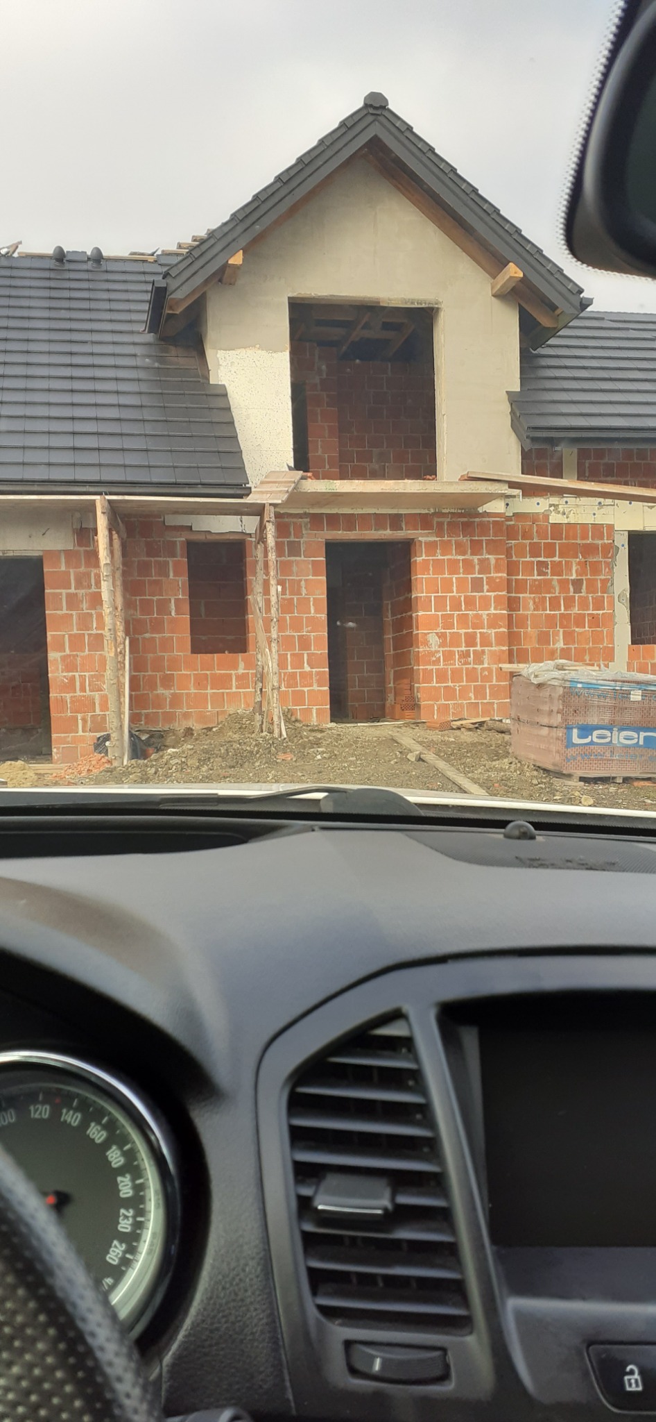 Surowy stan budowy domu z czerwonej cegły widziany z wnętrza samochodu, widoczne niezamknięte okna i drzwi, na dachu czarna dachówka.